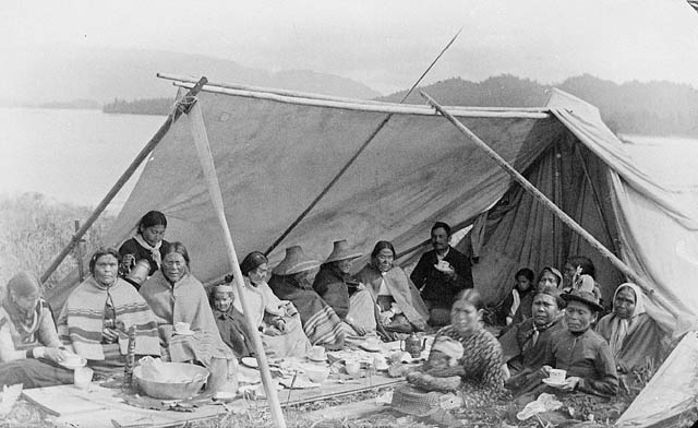 A group of people in historic clothing, some with traditional shawls, eat under a tent.