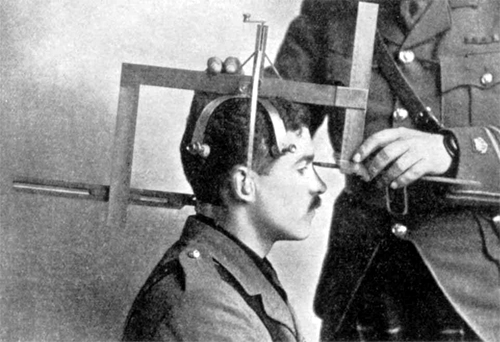 A standing person in uniform is using a large metal tool to measure a seated man’s head.