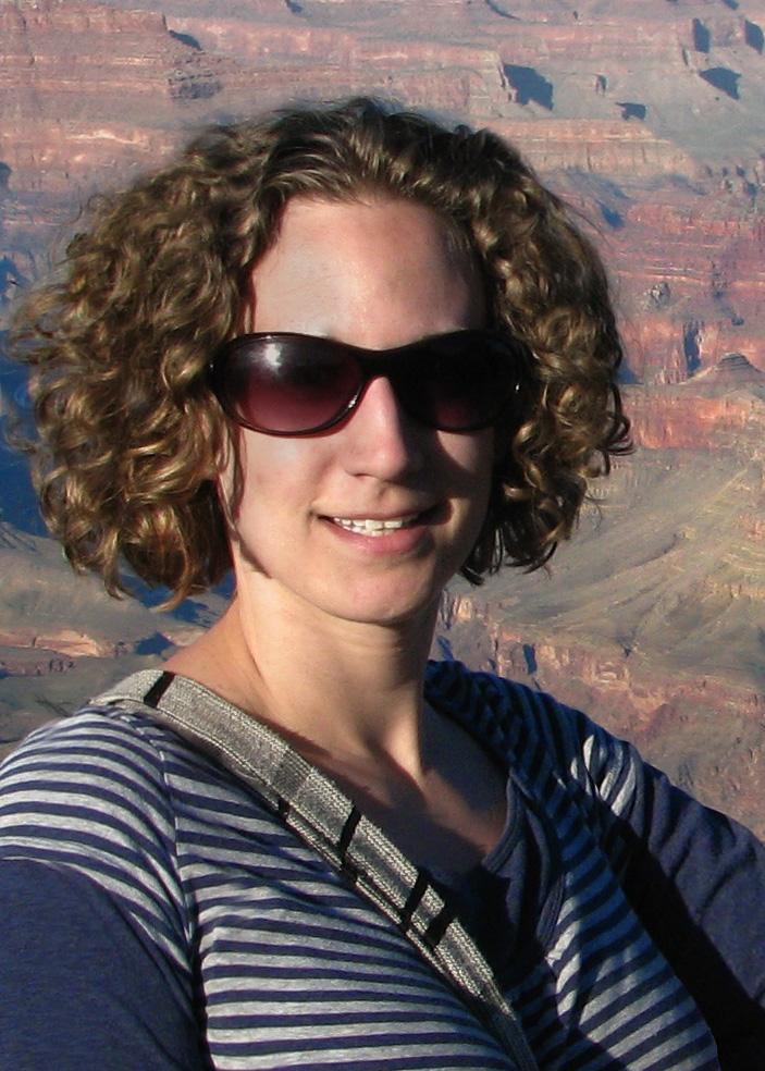 A woman with curly blonde hair and sunglasses smiles in front of a mountain landscape.