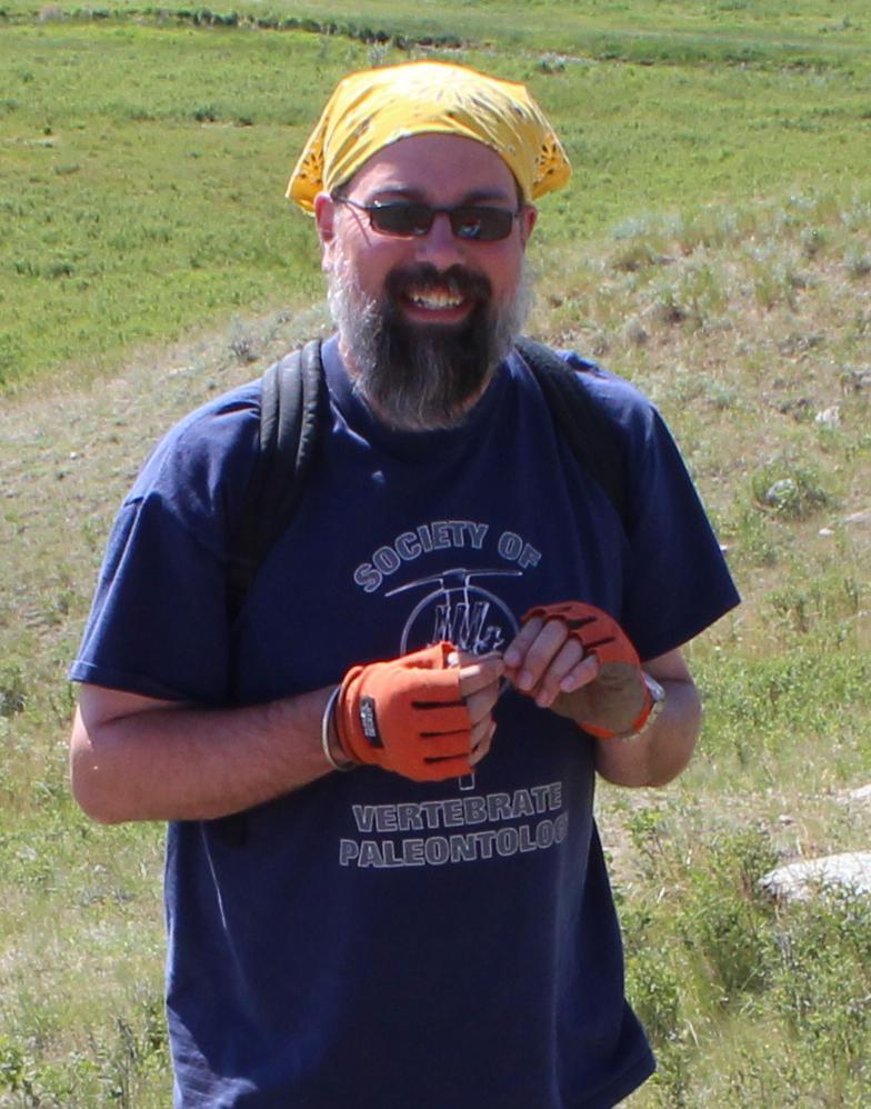 A man with sunglasses, a full beard, and a bandana stands in a field.