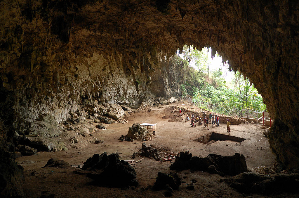 View from inside a large cave with people standing near a dug-out square of dirt.